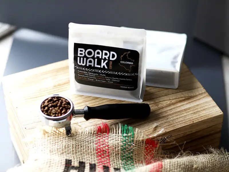 A Boardwalk Colombia bag on a wooden block beside a portafilter of beans and a coffee sack.