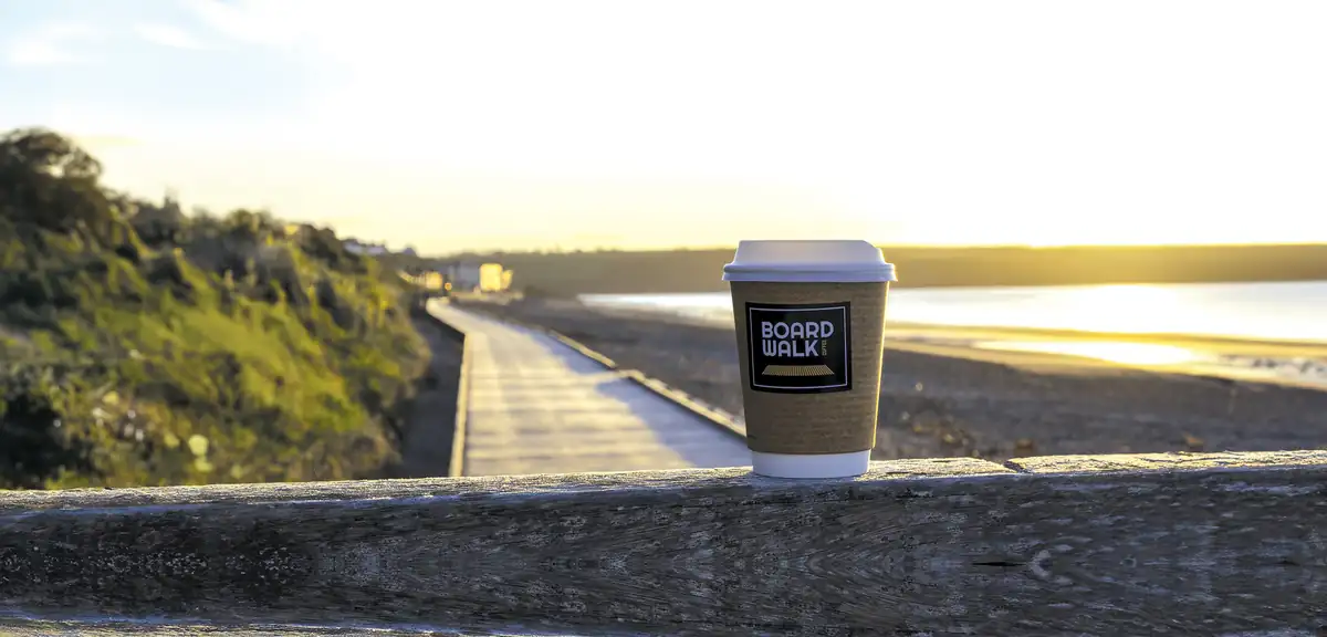 A Boardwalk takeaway cup on a wooden rail above the boardwalk and beach at Youghal, low sun behind.